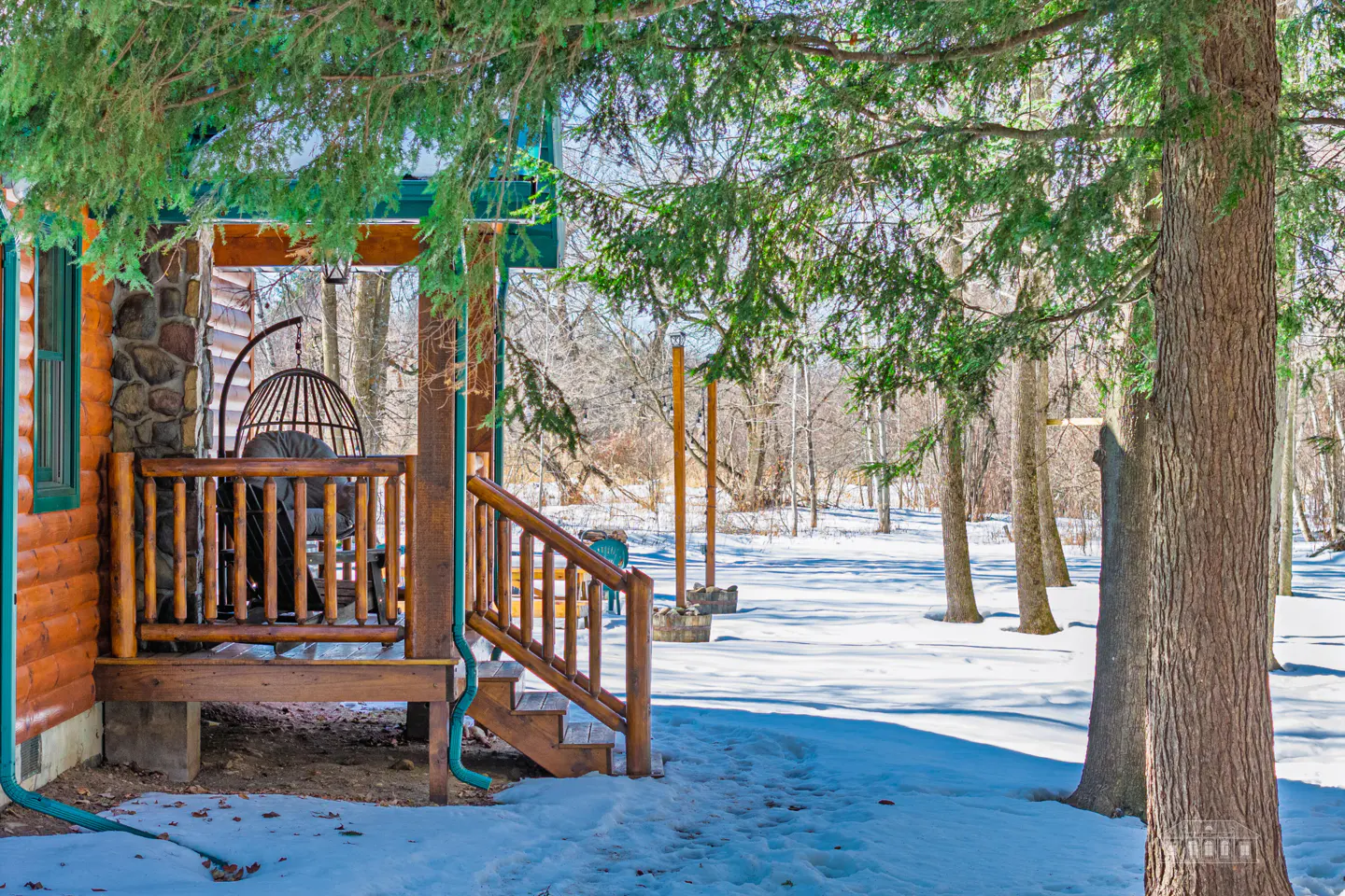 Log cabin porch with a hanging chair, wooden railings, and steps. Snow covers the ground, with trees in the background.