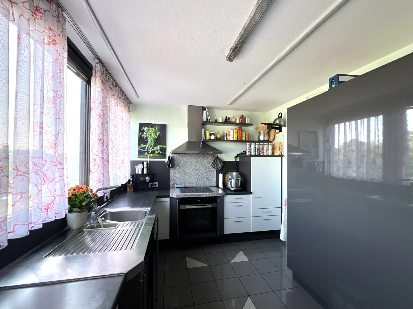 Modern kitchen with stainless steel sink, dark countertops, white cabinets, and a large grey refrigerator on tiled floors.