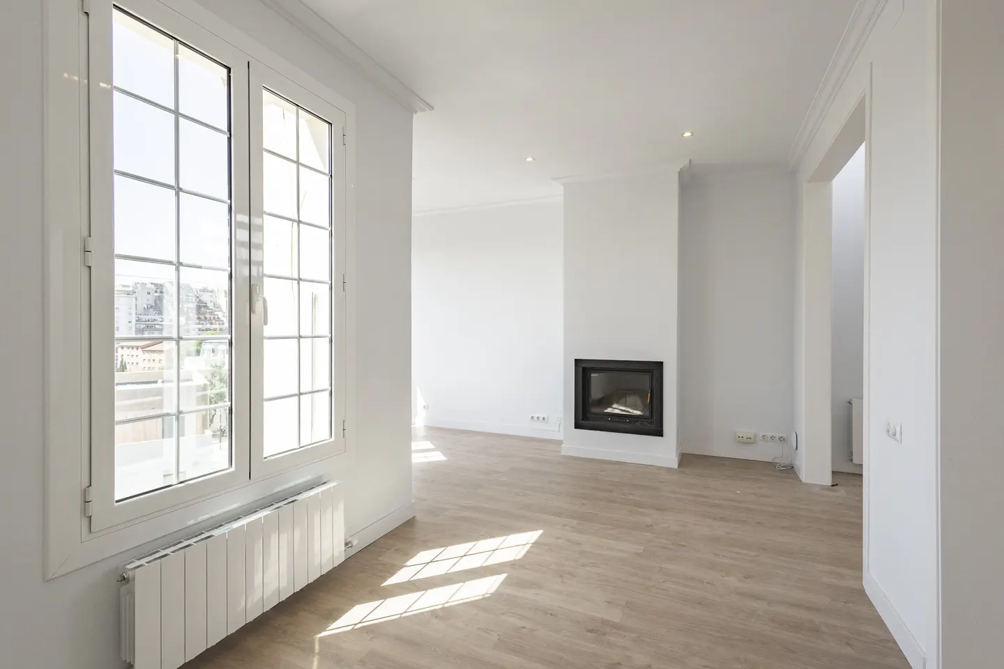 Bright, empty room with light wood floors, white walls, and a black fireplace. Large window with a radiator below.