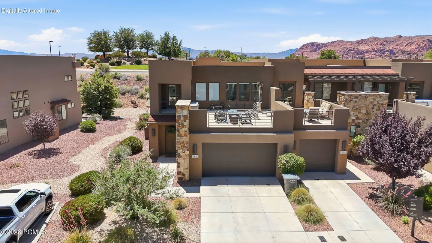 Modern two-story brown stucco home with a stone-accented balcony, two-car garage, and desert landscaping under blue skies.