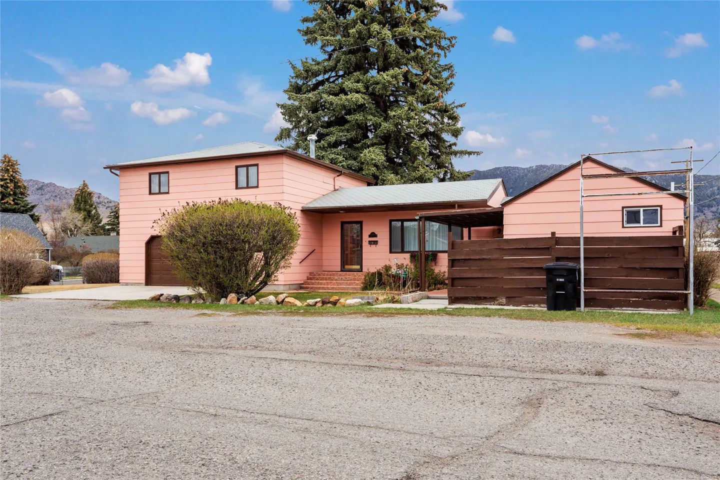 A two-story pink house with a brown fence, a large evergreen tree, and mountain views under a blue sky.