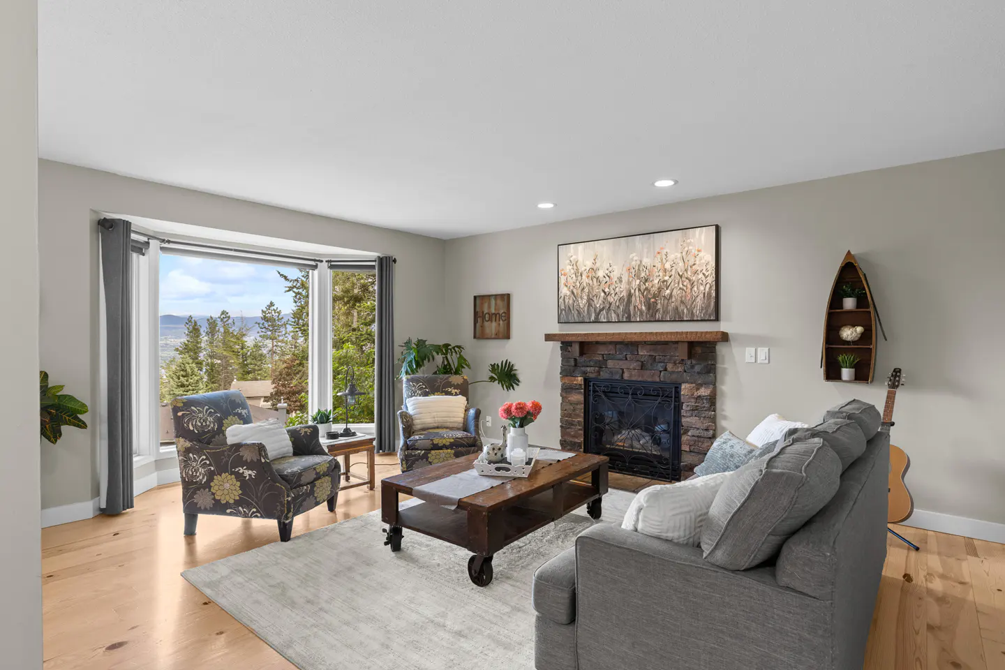 Living room with a gray sofa, floral armchairs, and a stone fireplace. A large window offers a view of trees and distant hills.