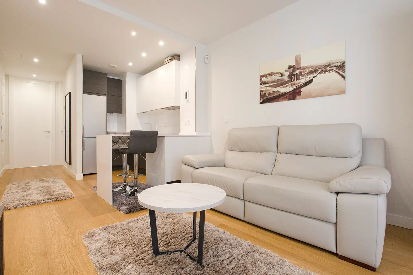 Bright apartment interior with a white sofa, round table, and kitchen bar with a gray stool. A city print hangs on the wall.