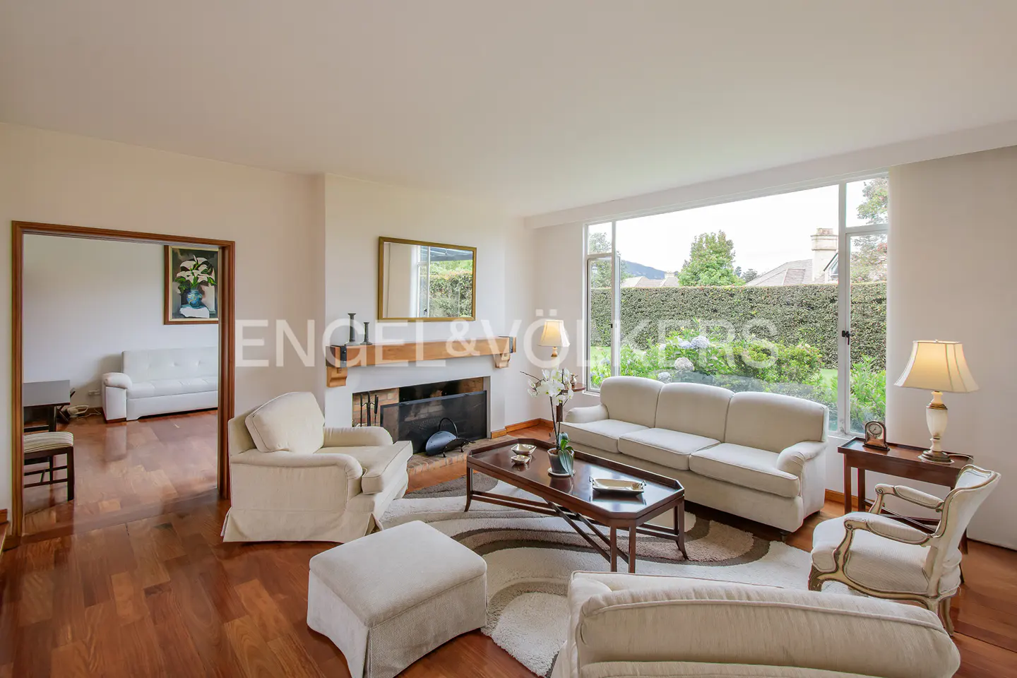 A bright living room with white sofas, chairs, and a fireplace. A large window overlooks a green garden.