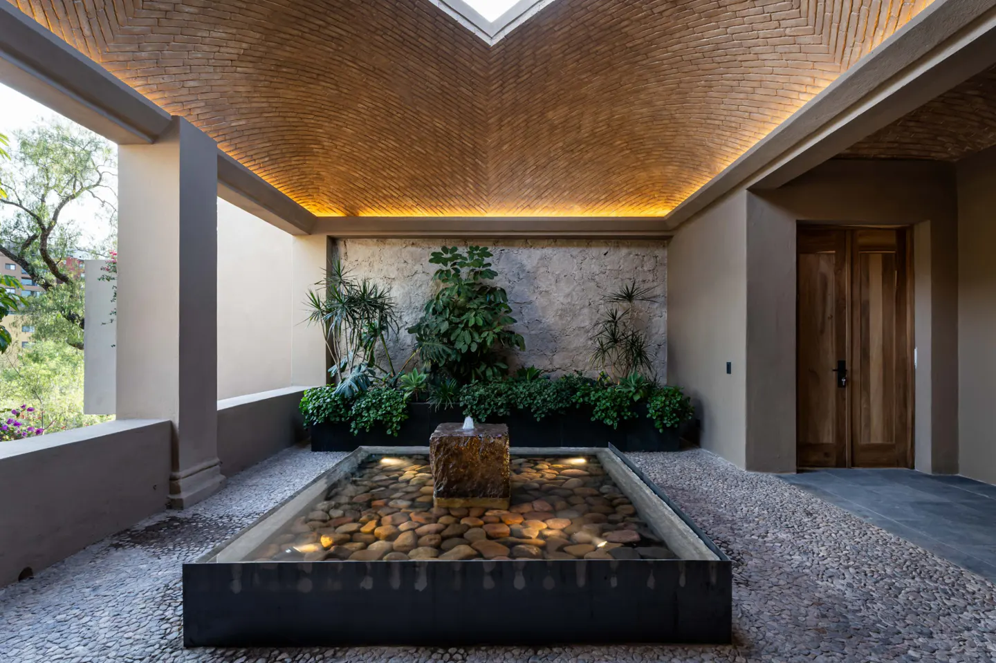 A courtyard with a brick vaulted ceiling, stone wall with plants, and a fountain filled with pebbles.