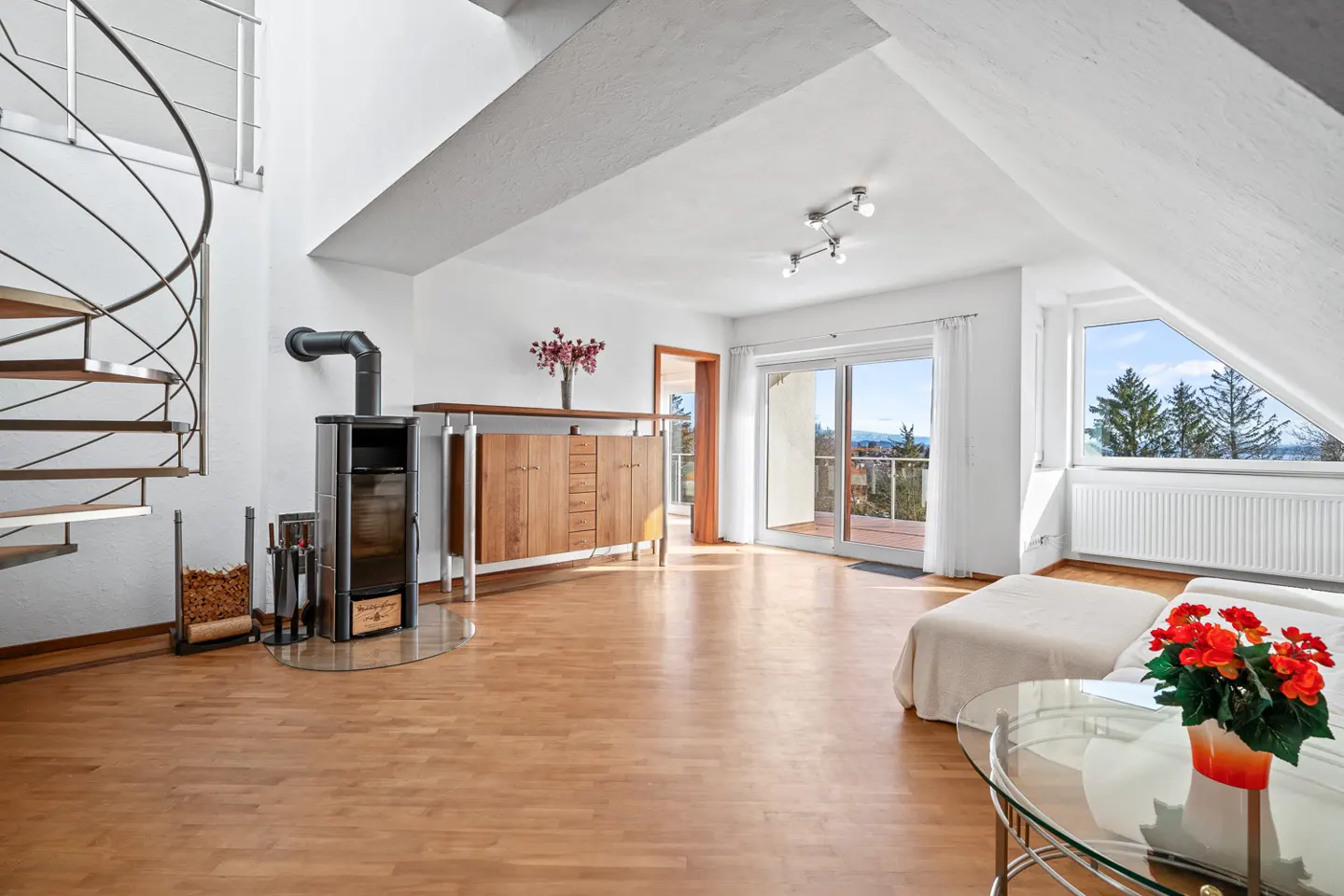 Bright, modern living room with wood floors, spiral staircase, wood-burning stove, and large windows with a view.