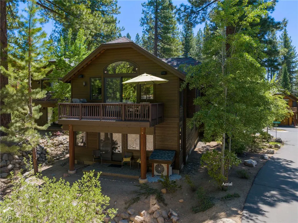 A two-story brown house with a balcony and a green umbrella, surrounded by trees.