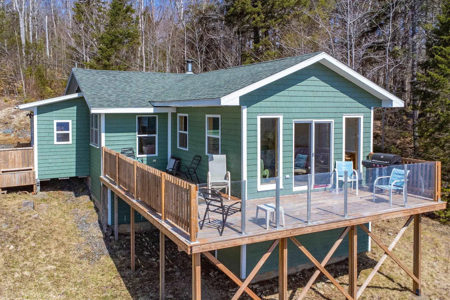 Exterior view of a green house with a deck, chairs, and a grill, surrounded by trees.