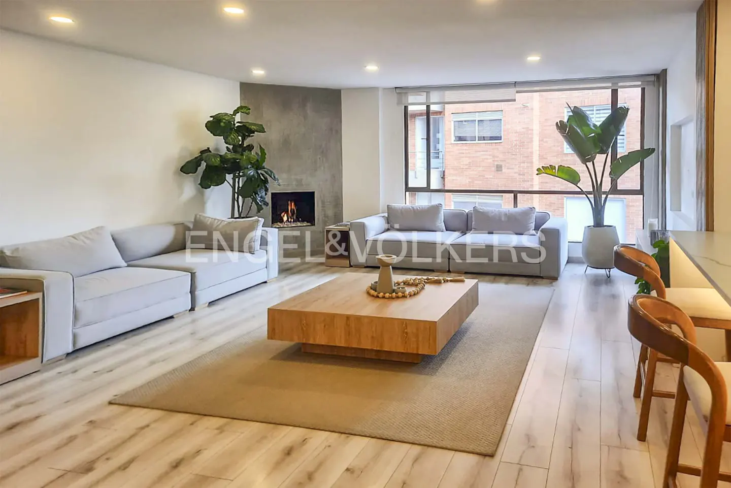 Modern living room with light wood floors, two grey sofas, a large wooden coffee table, and a fireplace with a plant.