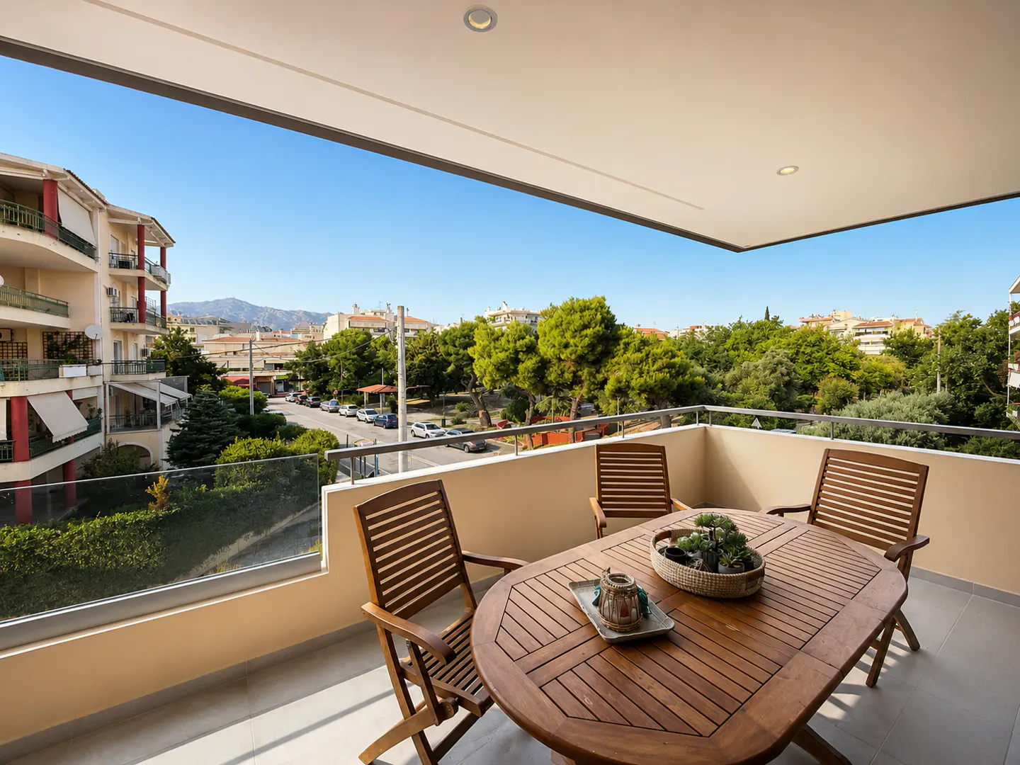 Covered balcony with a wooden table and chairs overlooking a sunny street, trees, and distant mountains.