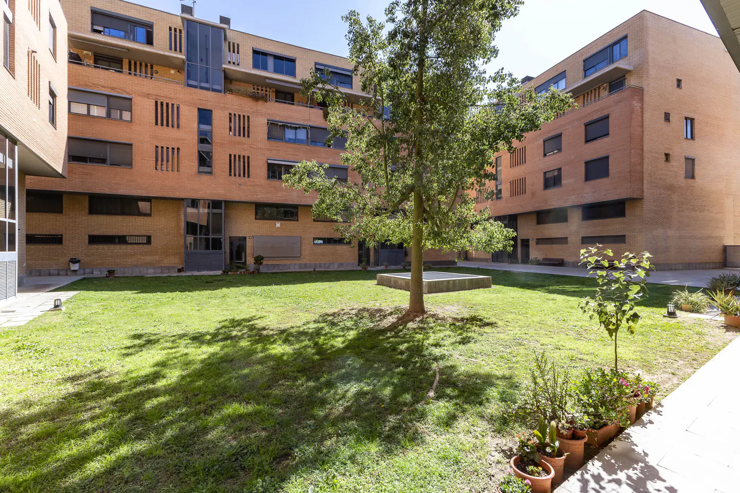 Courtyard view of a modern brick apartment building with a green lawn and trees. Potted plants line the walkway.