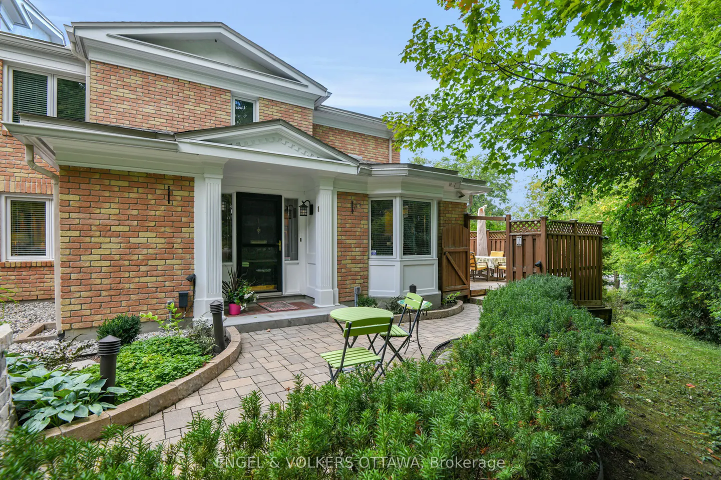 Exterior of a two-story brick house with a white-columned porch and a green bistro set on the stone walkway. A wooden deck is visible in the background.