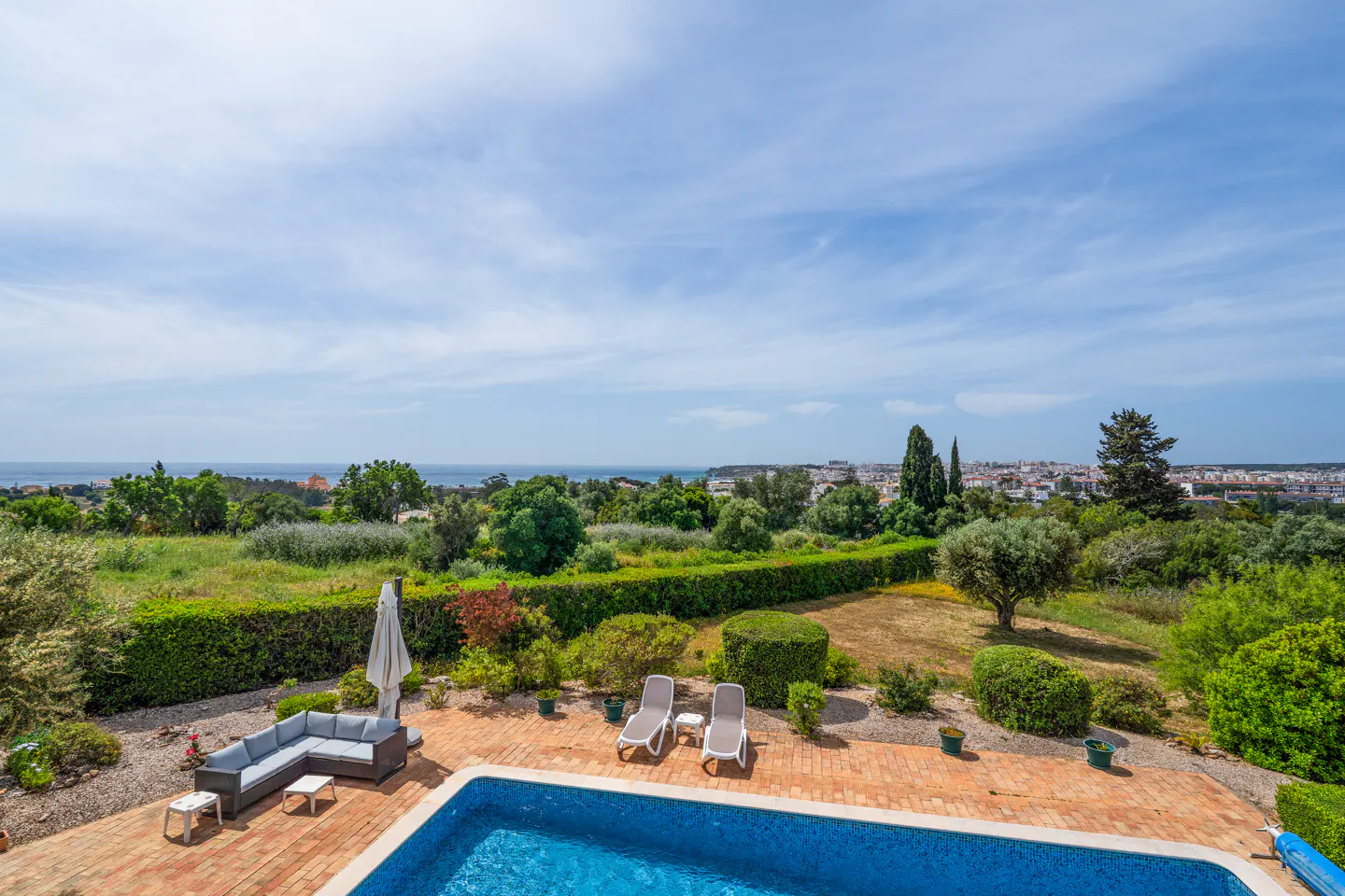 Aerial view of a blue pool, lounge chairs, and a gray sofa on a brick patio, with green trees, a town, and the ocean in the background.