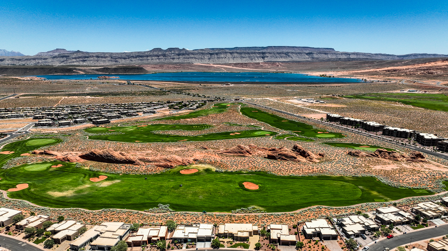 Aerial view of a golf course community with green fairways, red rock formations, homes, a lake, and mountains under a blue sky.
