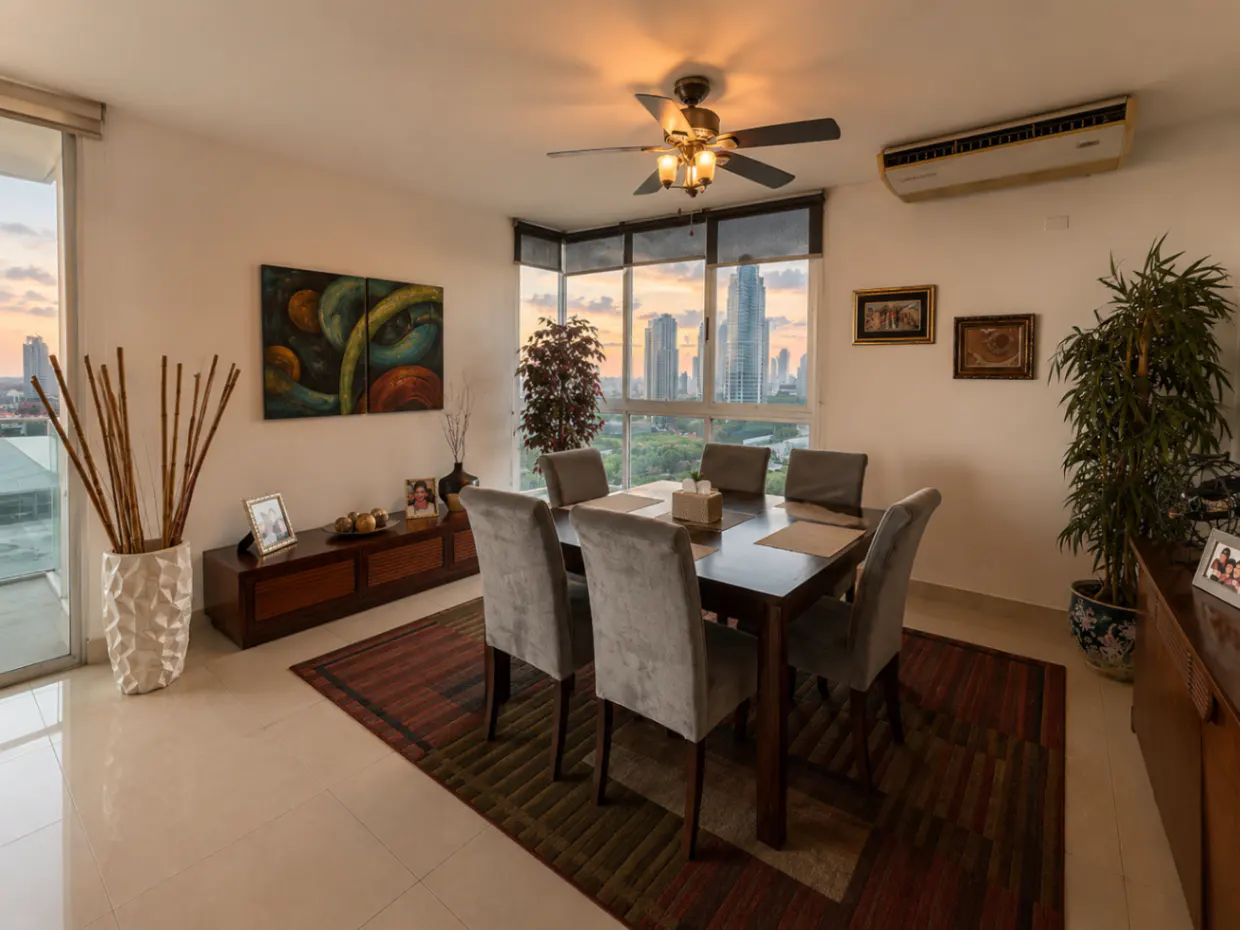 Bright dining room with a dark wood table, six gray chairs, and a city view through large windows. A ceiling fan and artwork add detail.