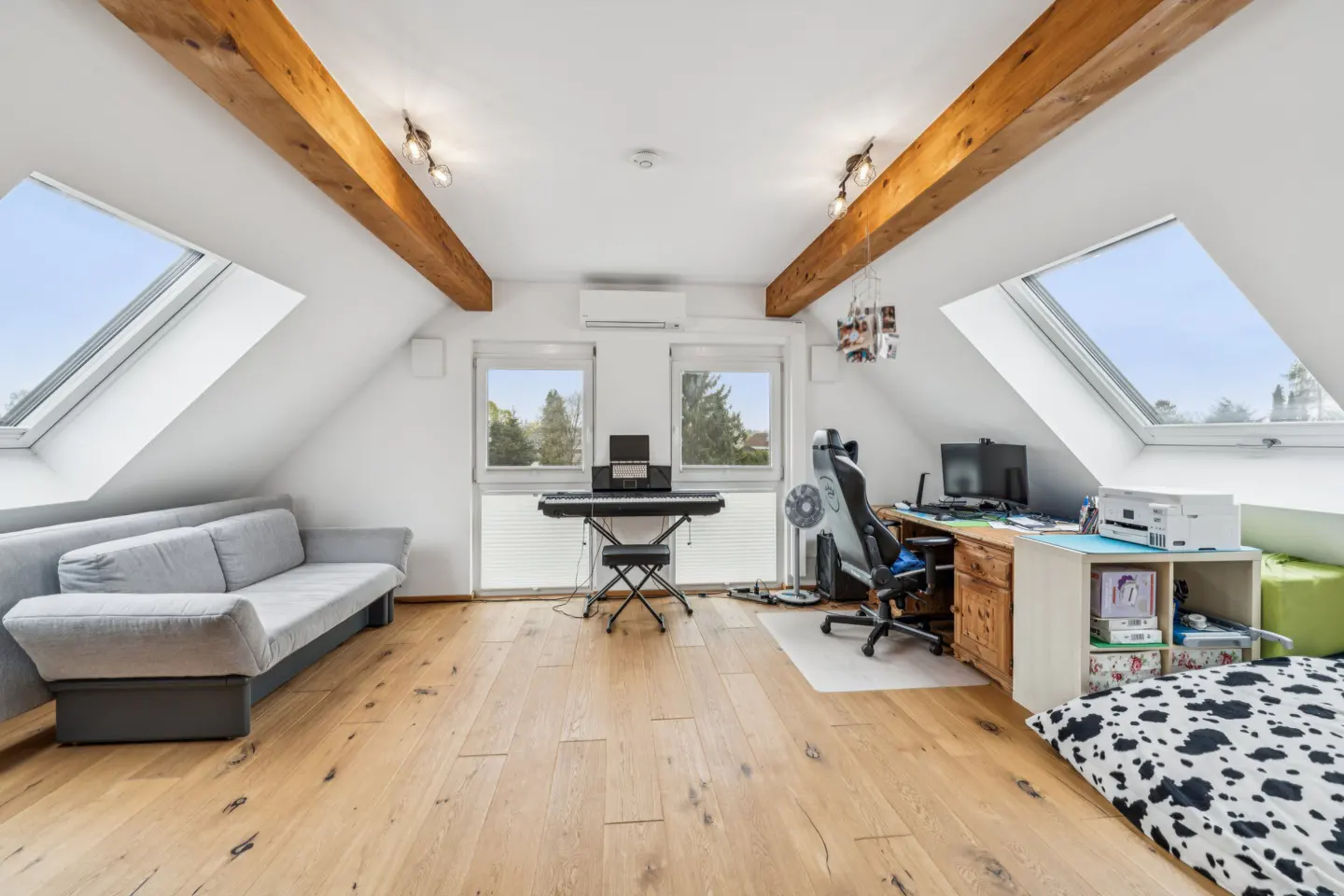 Attic room with wooden beams, skylights, and hardwood floors. A gray sofa, keyboard, desk, and chair furnish the space.