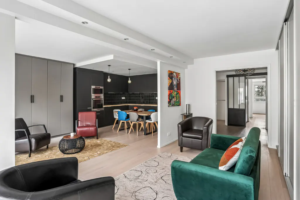Open-concept living space with a green sofa, black chairs, and a dining area with colorful chairs. The kitchen has black cabinets and stainless steel appliances.