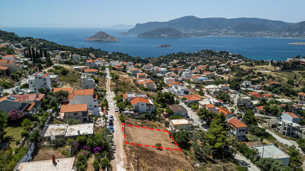 Aerial view of a coastal town with a vacant plot outlined in red, surrounded by houses, hills, and a blue sea with islands.