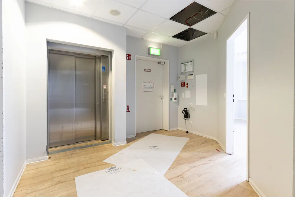 Hallway with a silver elevator, a white door with a green exit sign, and light wood floors covered with white protective sheets.