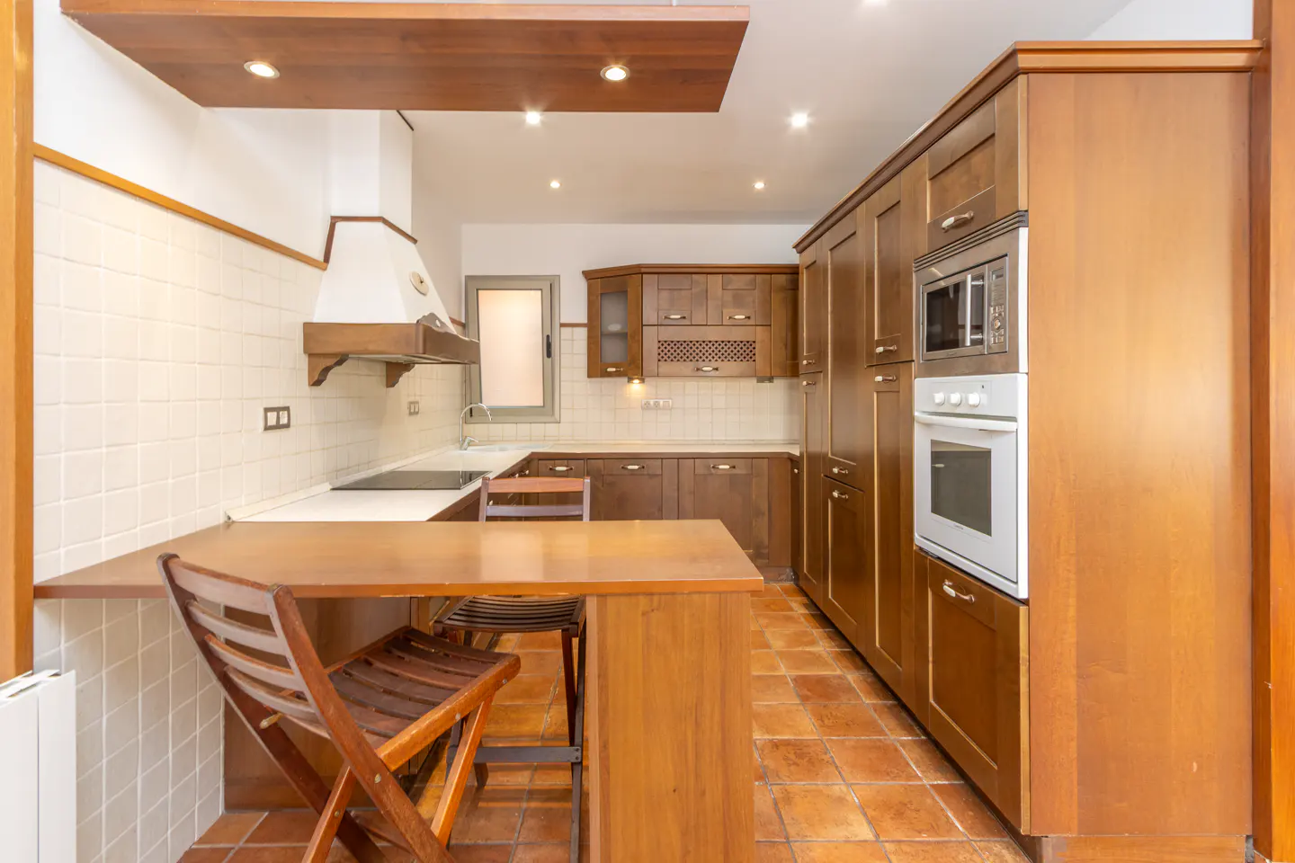 A kitchen with wooden cabinets, a white oven, a microwave, and a wooden table with chairs. The floor is tiled in terracotta.