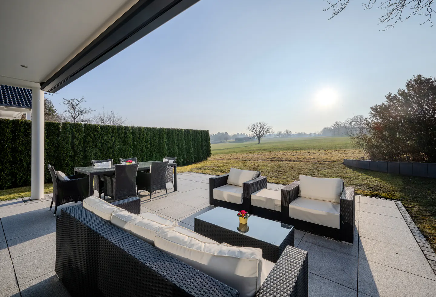 Outdoor patio with black wicker furniture, white cushions, glass table, and dining set overlooking a green lawn and trees on a sunny day.