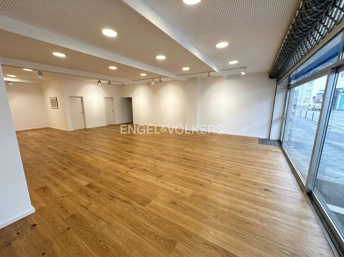 Empty retail space with light wood floors, white walls, and a perforated white ceiling with recessed lighting. Large windows face the street.