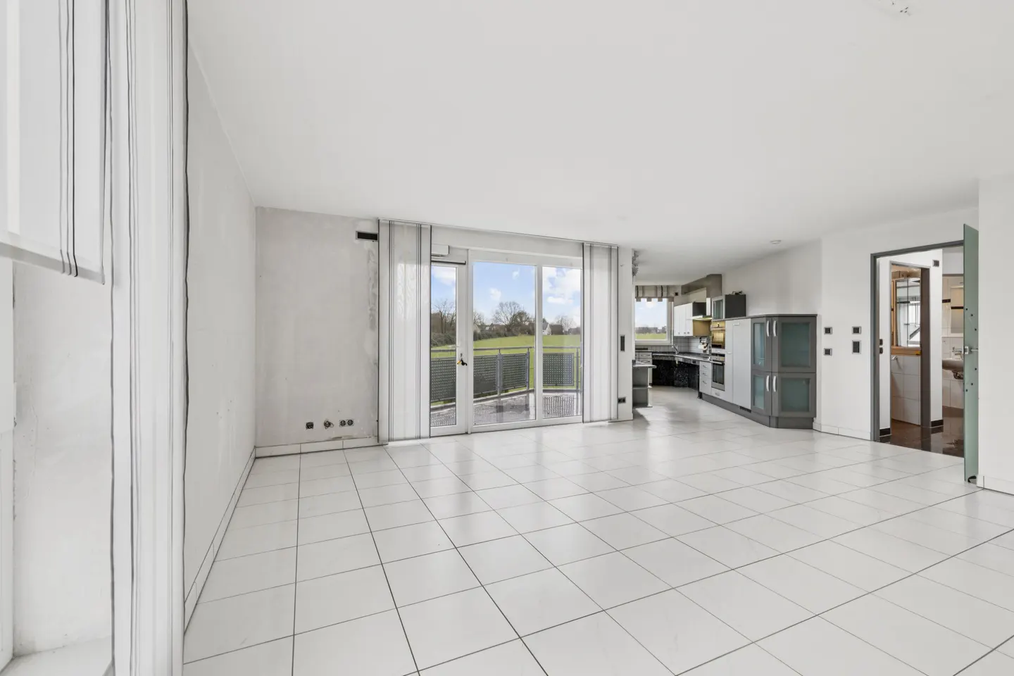 Bright, empty room with white tile floor, white walls, and sliding glass doors to a balcony overlooking a green field. Kitchen area visible in the background.