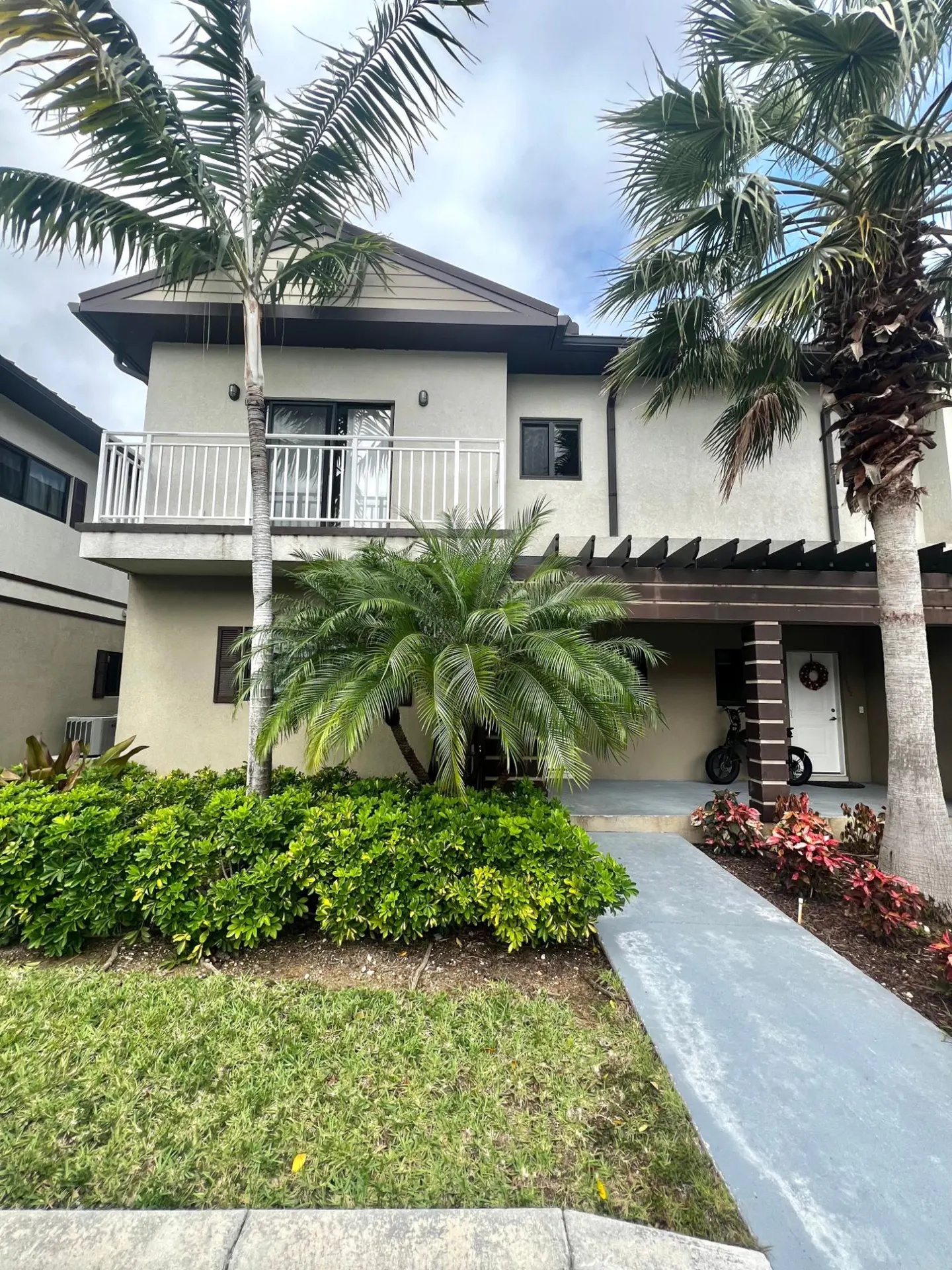 Two-story tan townhouse with a balcony, brown trim, and a pergola over the front door. Palm trees and green bushes surround the building.