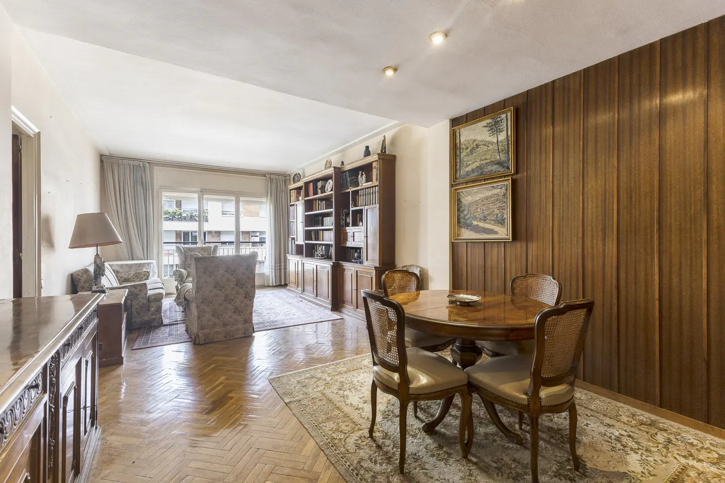A living and dining room with wood paneled walls, a round table with four chairs, and a bookshelf.