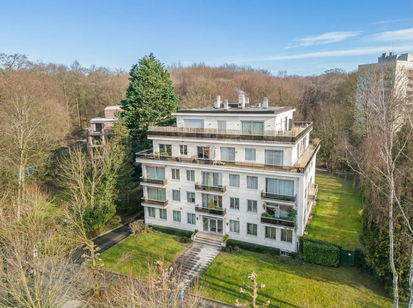 A four-story white apartment building with balconies, surrounded by green lawns and trees under a blue sky.