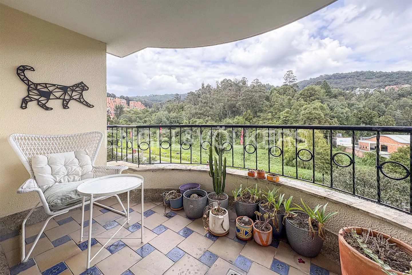 Balcony with a white wicker chair, table, and potted plants. A geometric cat wall decoration is visible. The view overlooks green hills.