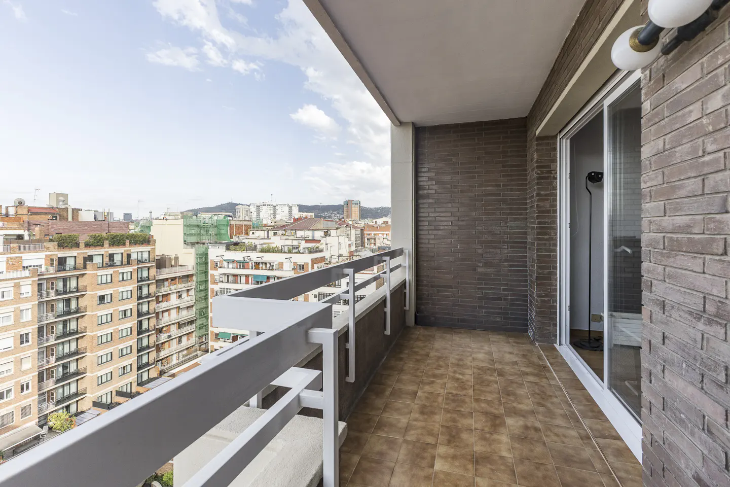 Balcony view with white railings overlooking a cityscape under a cloudy sky. Brown brick wall and tiled floor. Sliding glass door to the right.