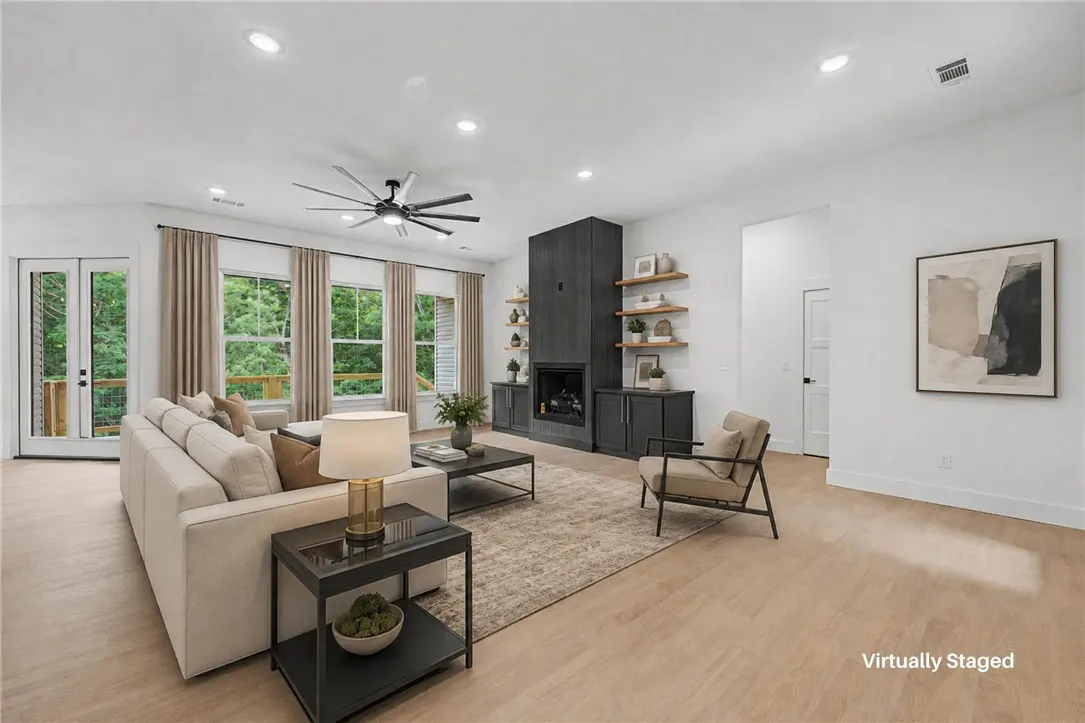 Bright living room with a white sofa, black fireplace, and light wood floors. Windows with beige curtains overlook a green outdoor space.