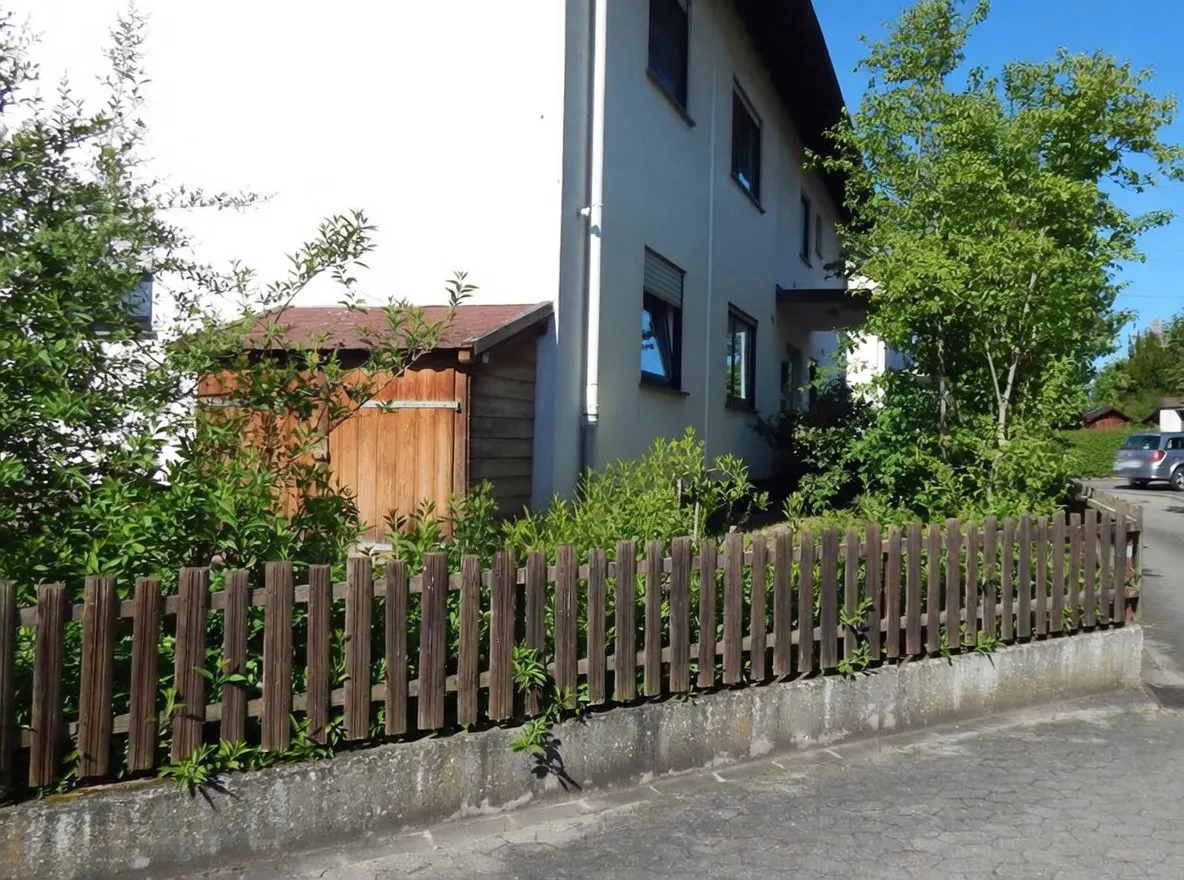 A two-story white house with a brown wooden fence and shed, surrounded by green trees and bushes.
