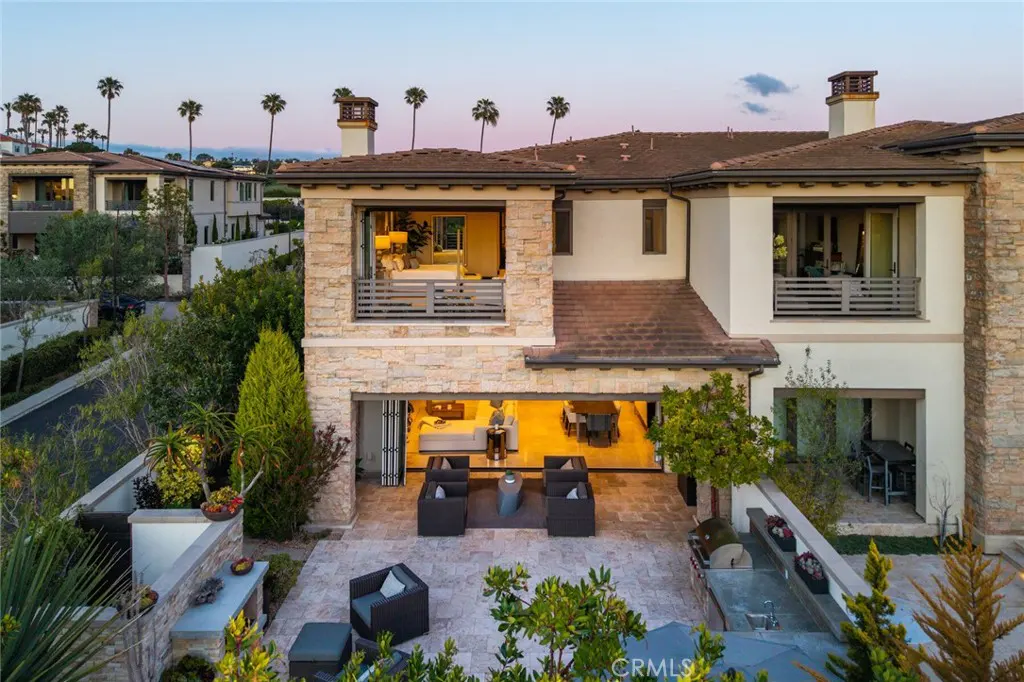 Exterior view of a two-story house with a stone facade, open patio, and palm trees in the background.