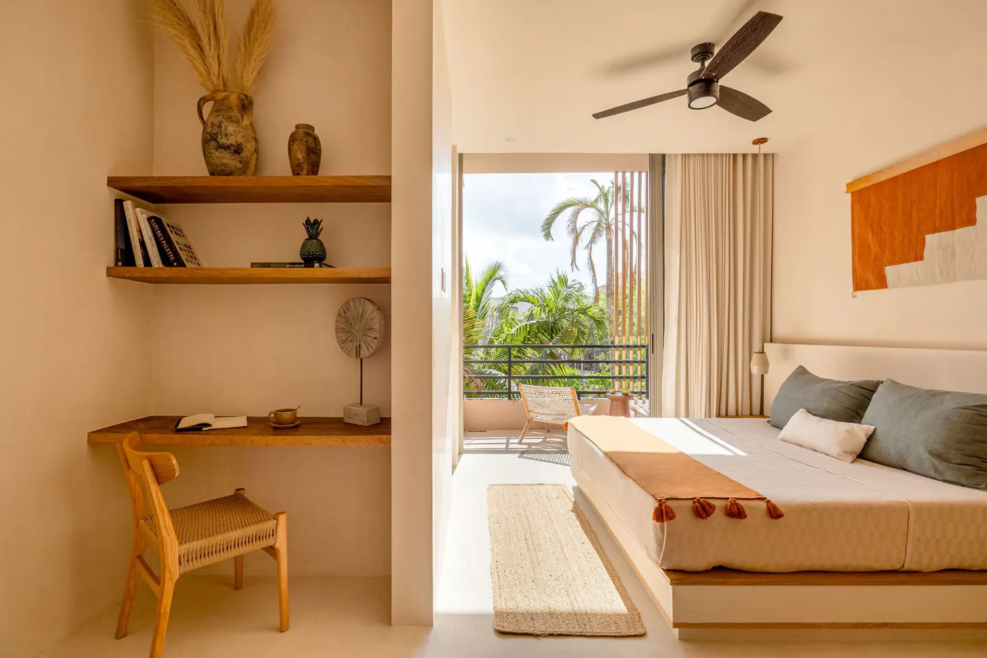 Bedroom with a desk, shelves, and a balcony with tropical plants. The bed has gray pillows and an orange throw. A ceiling fan is visible.