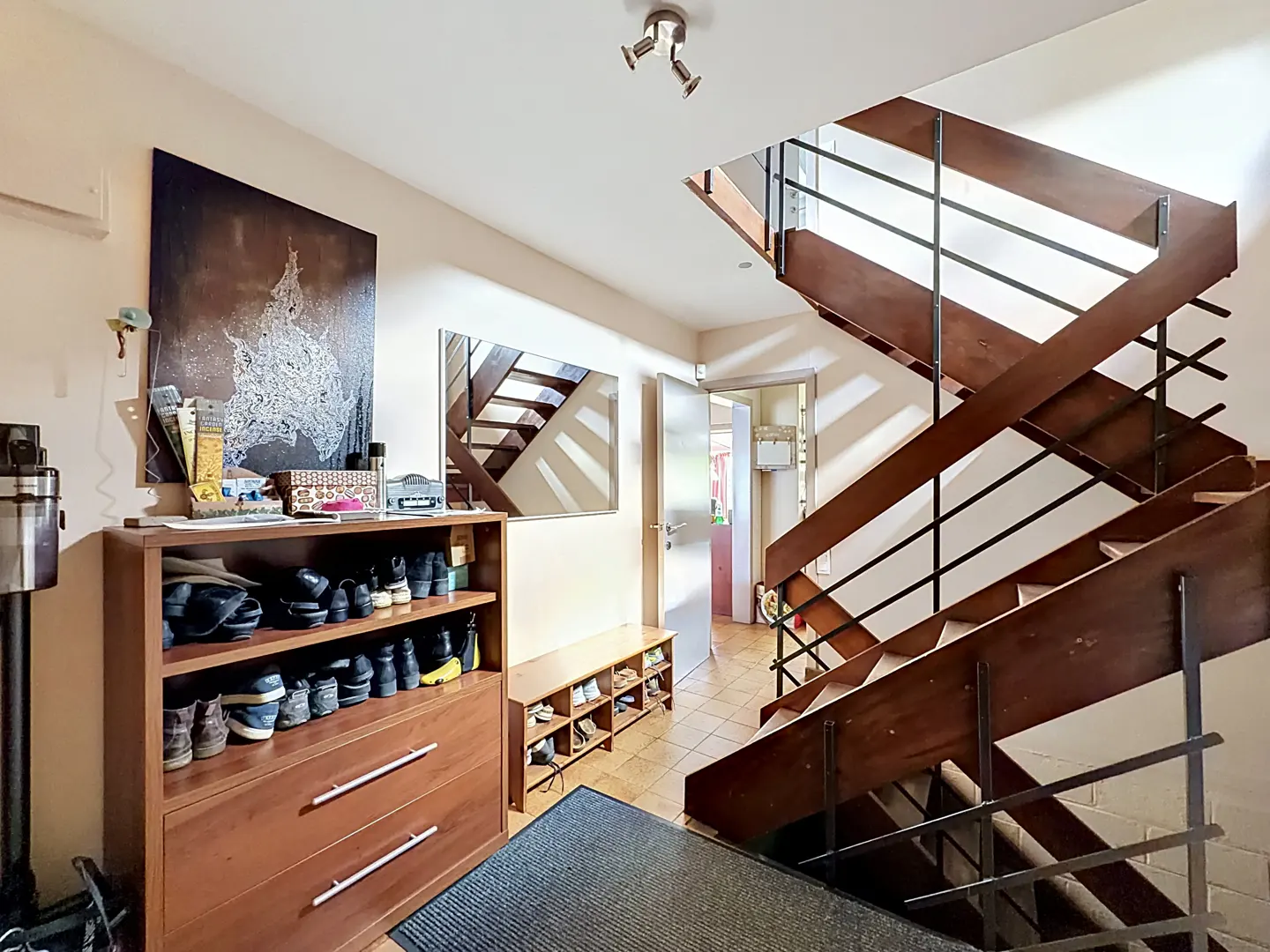 Entryway with a wooden shoe rack, art, and a staircase with black railings. A mirror reflects the stairs.