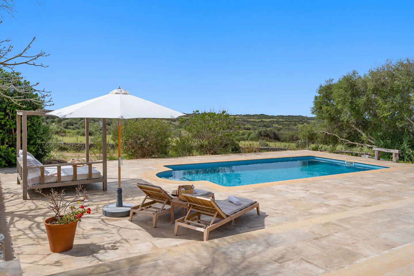 Outdoor pool area with lounge chairs, daybed, and umbrella on a stone patio. Green trees and a clear blue sky in the background.