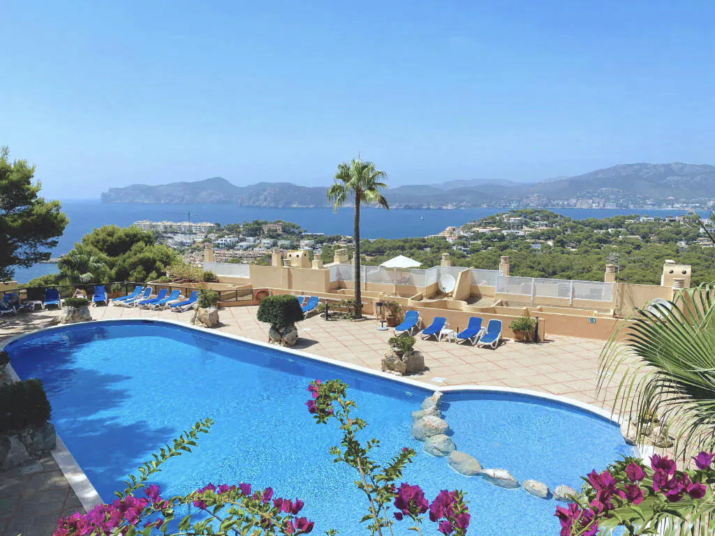 Scenic view of a blue swimming pool with lounge chairs, overlooking the ocean and green hills under a clear blue sky.