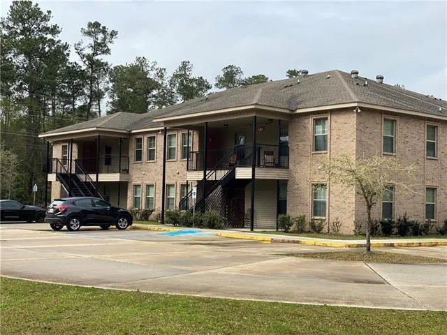 Two-story tan brick apartment building with exterior staircases, a parking lot, and trees under a cloudy sky.