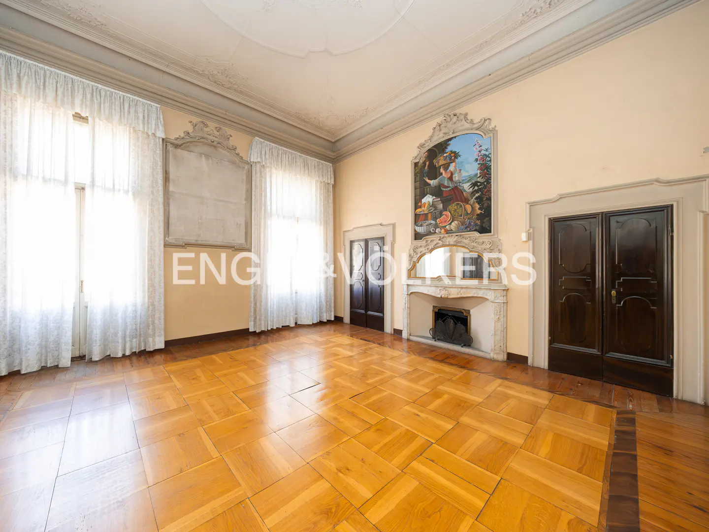 A spacious room with parquet floors, tall windows with white curtains, and a decorative fireplace with a painting above it.