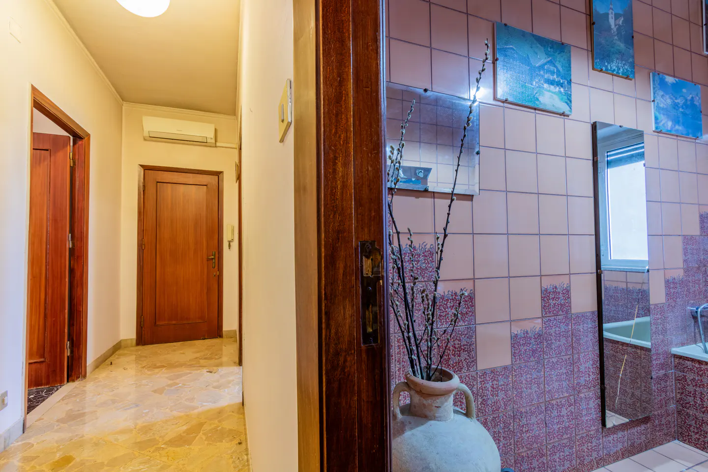 Interior view of a hallway with marble floors, wooden doors, and a tiled bathroom with mirrors and artwork.
