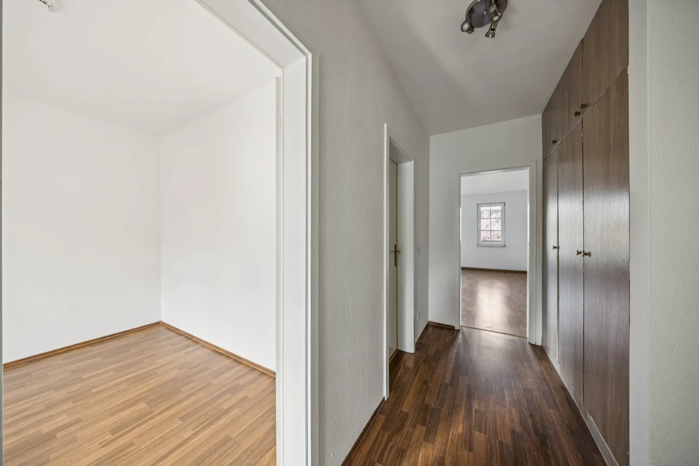 Hallway with wood floors, white walls, and built-in wood cabinets. A doorway leads to an empty room with wood floors.
