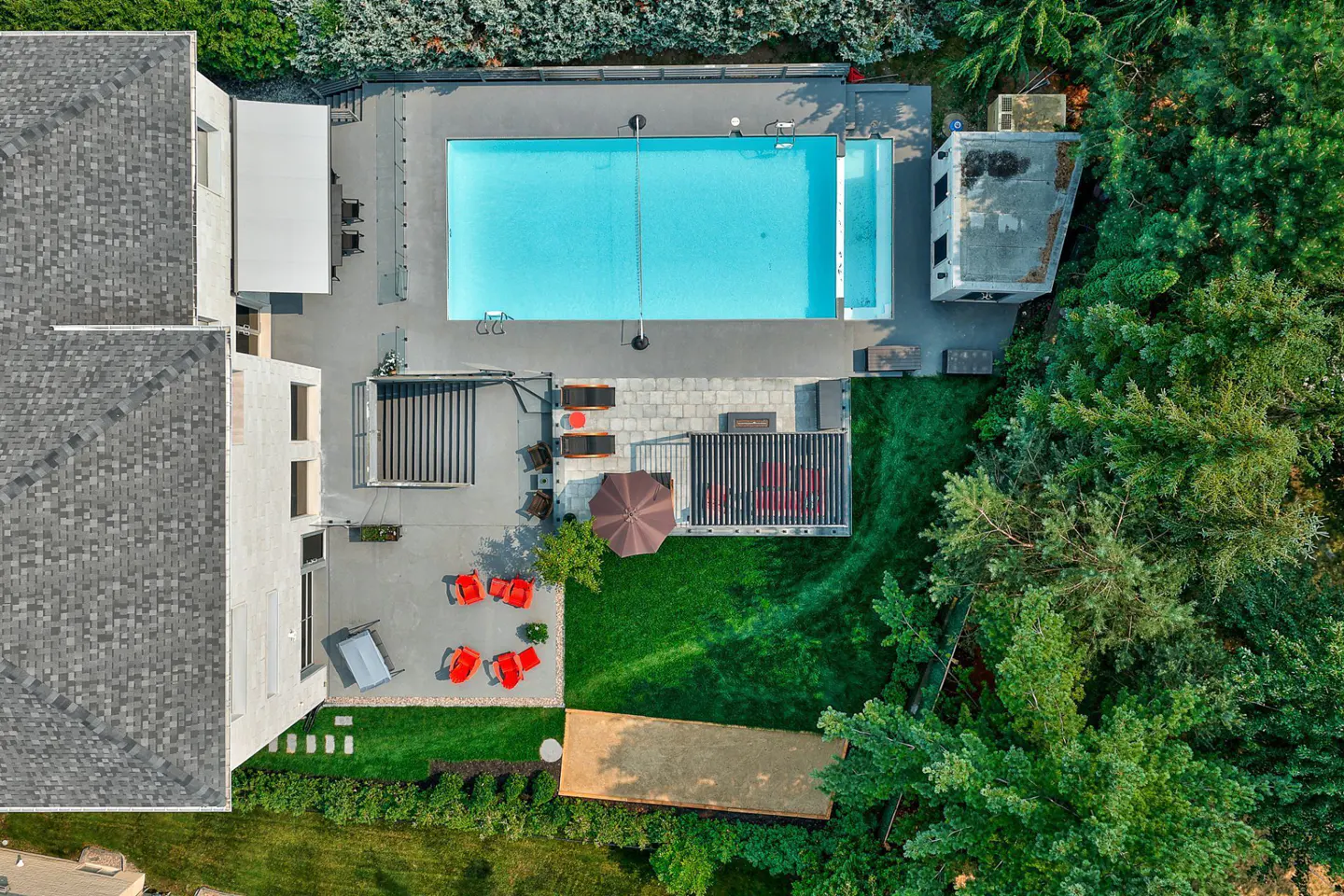 Aerial view of a modern home with a pool, patio, and lush green landscaping. Red chairs and umbrellas add pops of color.