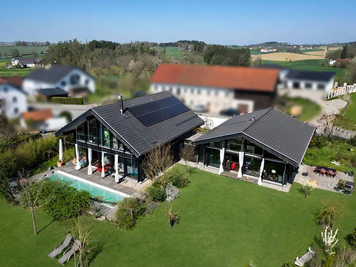 Aerial view of two modern, black-framed houses with gray roofs, solar panels, a pool, and a green lawn.