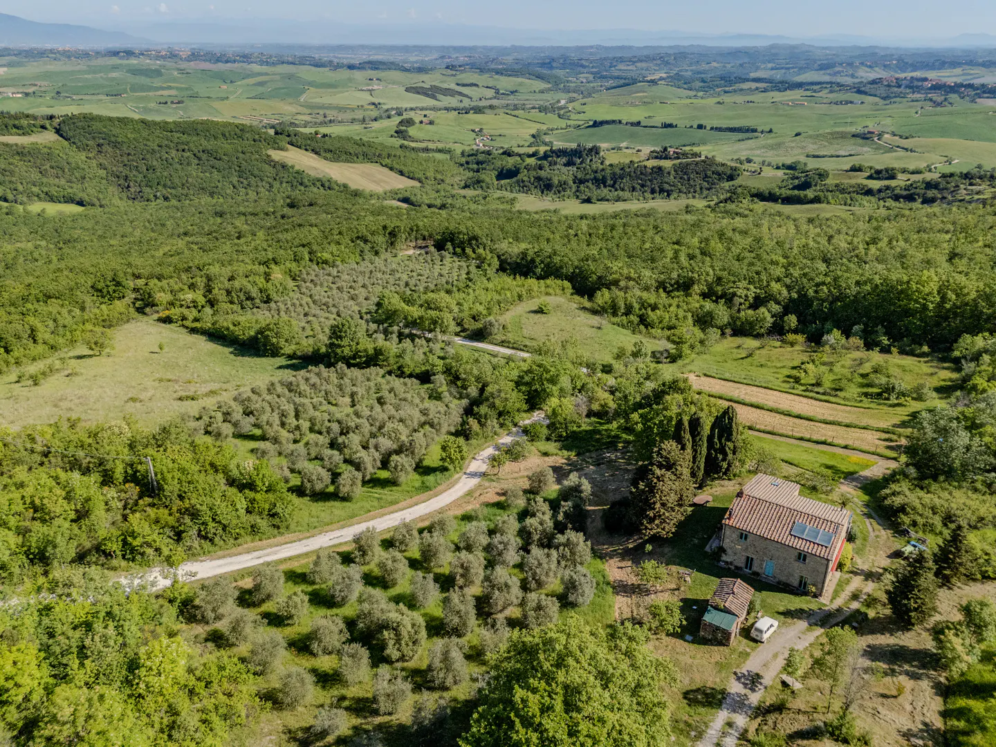 Aerial view of a stone house with a red tile roof, surrounded by green trees and fields in the Tuscan countryside.