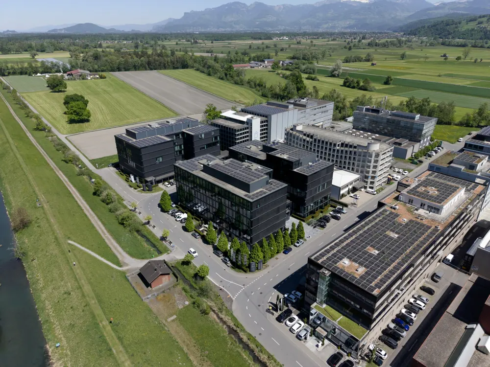 Aerial view of modern office buildings with solar panels, surrounded by green fields and mountains in the background.