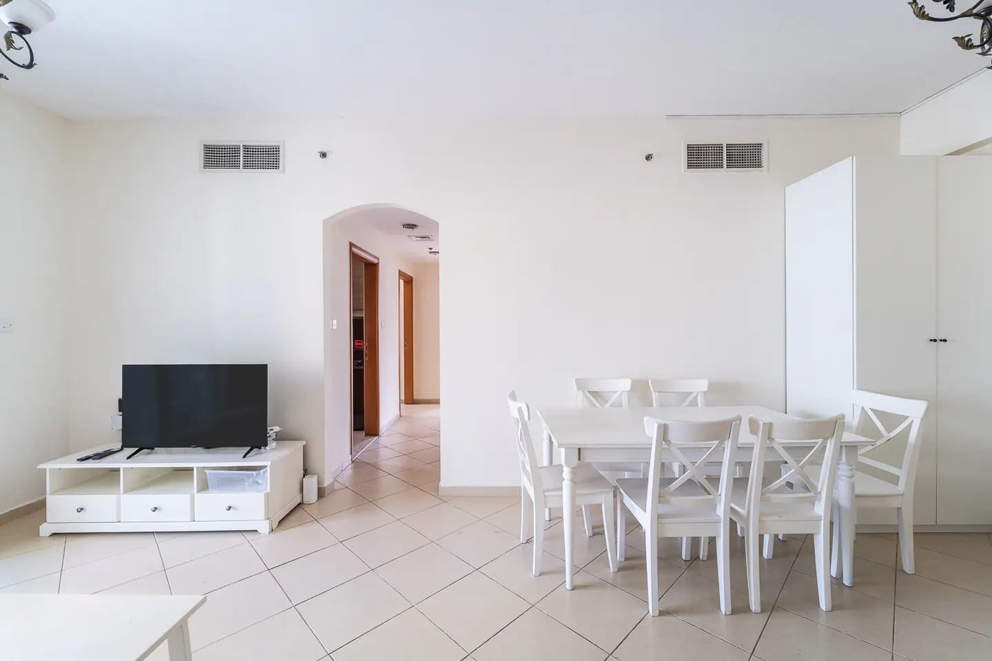 Bright, airy living room with white walls, tile floor, and a white dining set. A black TV sits on a white console table. Hallway visible through an archway.