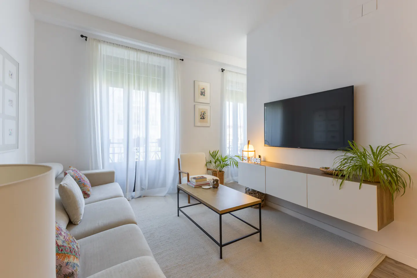 Bright living room with a beige sofa, a wooden coffee table, and a large TV on a white wall. Sheer white curtains filter sunlight.