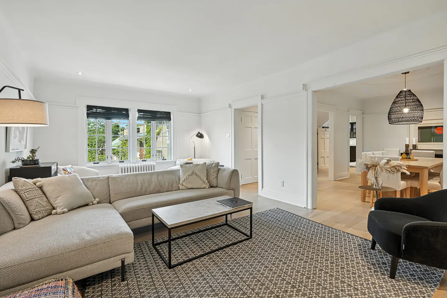 Bright living room with a beige sectional sofa, patterned rug, and a view into the dining room with a wooden table.